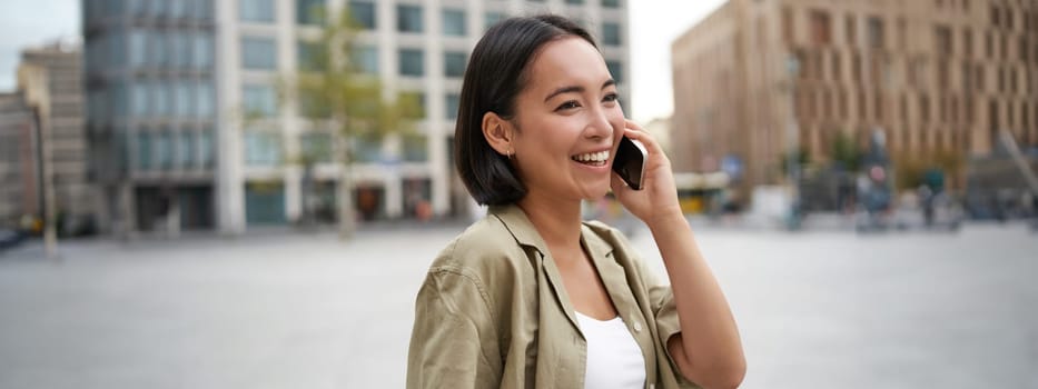 Modern young asian girl talks on mobile phone, uses telephone on city street. Woman smiling while calling someone on smartphone.