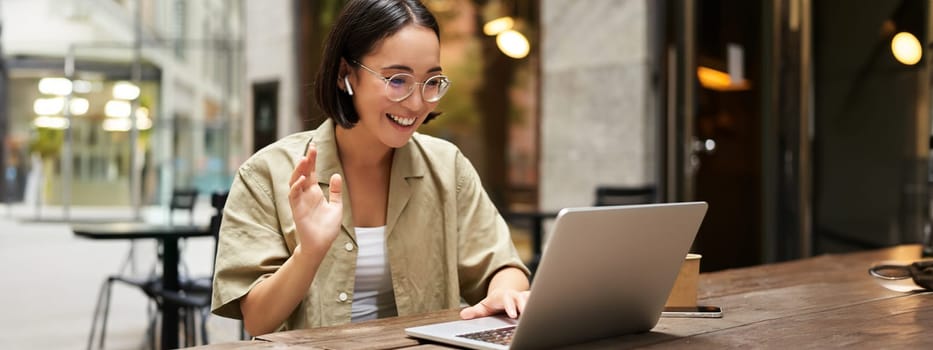 Online meeting. Young urban girl, asian woman talking with laptop, video chat, gesturing, sitting in an outdoor cafe and working remotely.