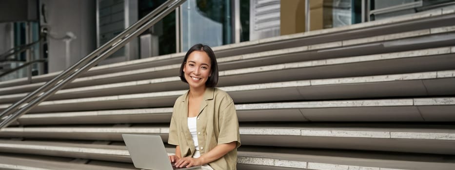 Vertical shot of asian girl sits with laptop, drinks coffee on university stairs. Young woman, student does her homework outdoors.