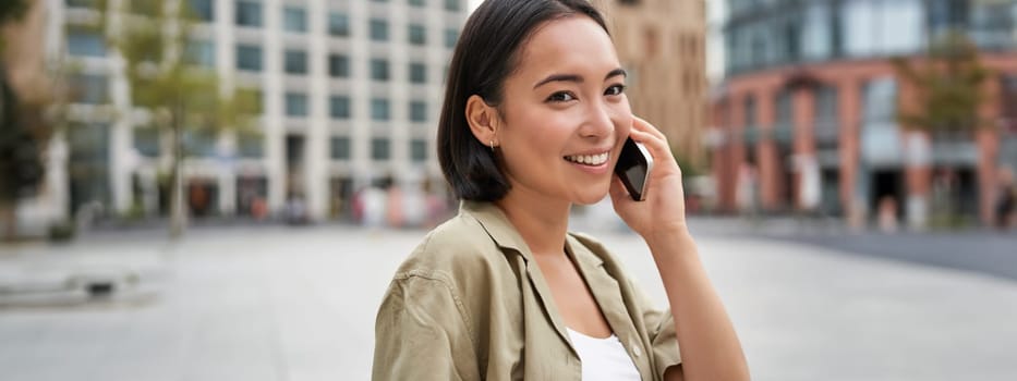 Cellular connection. Young asian woman makes a telephone call, talking on mobile smartphone and walking on street.