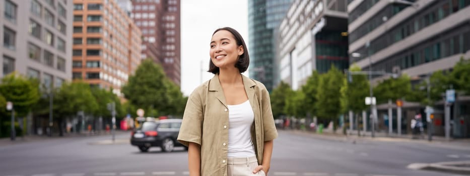 Outdoor shot of young smiling asian woman, standing on street in daytime, looking around, posing happy.