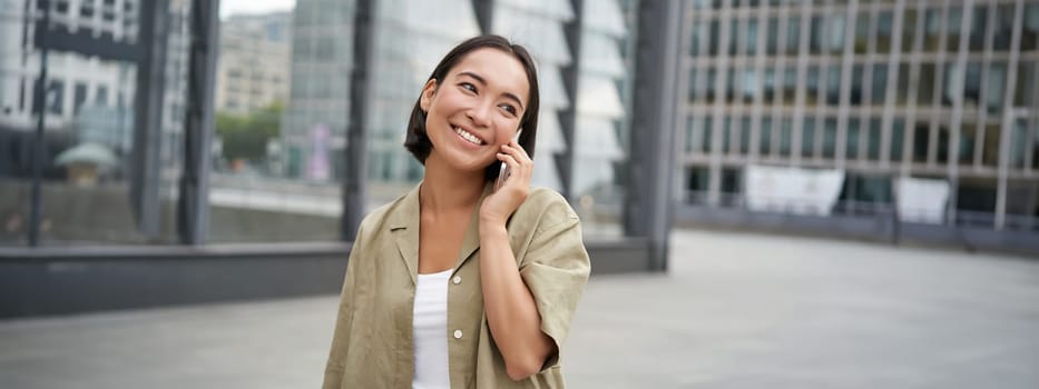 Smiling young korean girl talking on mobile phone and walking in city. Happy woman posing on street with smartphone.