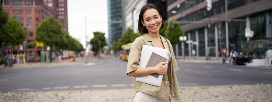 Portrait of young asian woman, looking happy and confident, going to work or university, city skyscrappers behind her, holding laptop and notebook.