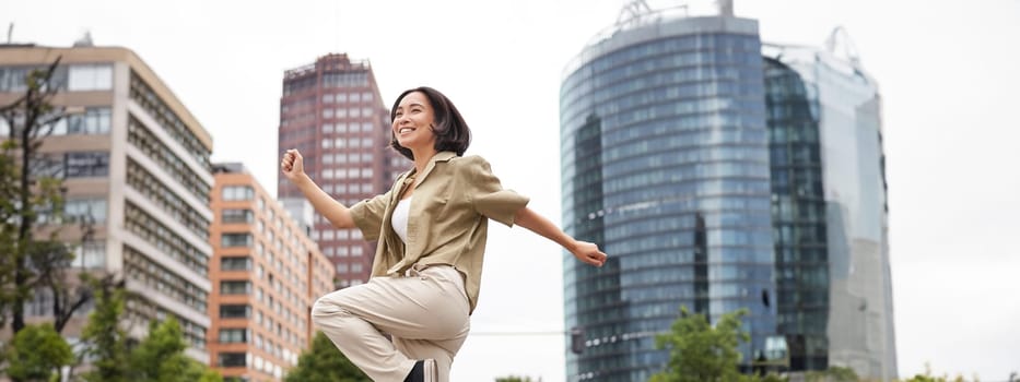 Vertical shot of young asian woman posing happy, raising hands up and dancing, triumphing, celebrating victory, enjoying day out in city.