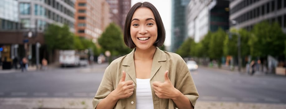 Enthusiastic city girl, shows thumbs up in approval, looking upbeat, say yes, approves and agrees, stands on street.