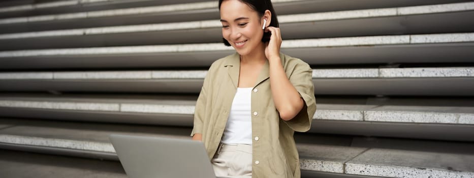 Remote worker. Smiling asian girl sits outdoors on street with laptop. Happy young woman working on computer remotely, listen music in wireless headphones.