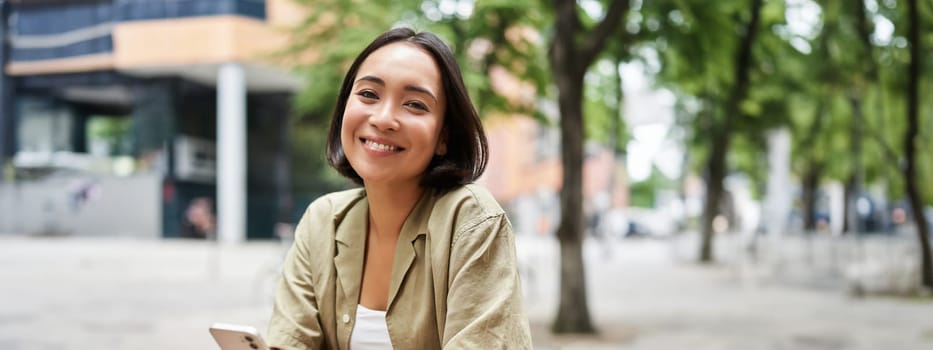 Portrait of young woman enjoying her coffee, drinking takeaway on bench in city, using smartphone. Copy space