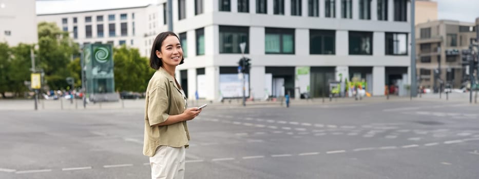 Car sharing and technology. Young asian woman waiting for taxi near road, holding smartphone, order car in application.