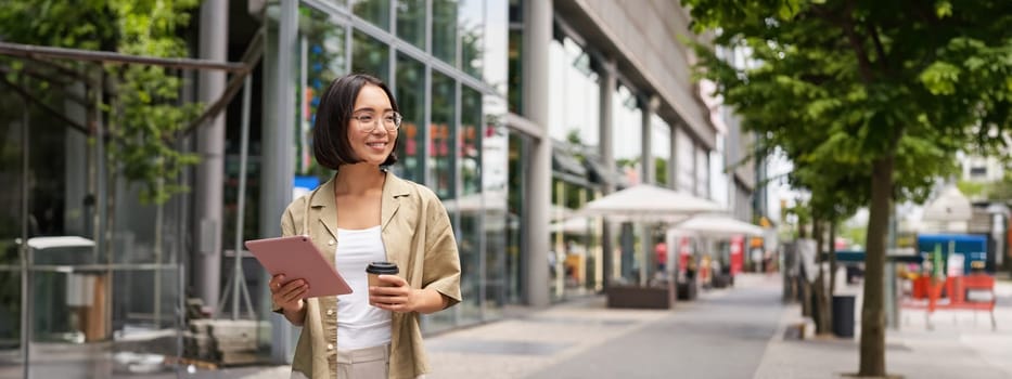 Smiling korean girl walking in city street with tablet, drinking takeaway coffee and looking happy.