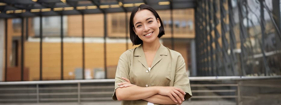 Portrait of young asian woman standing with confidence, cross arms on chest and smiling, posing outdoors on street.