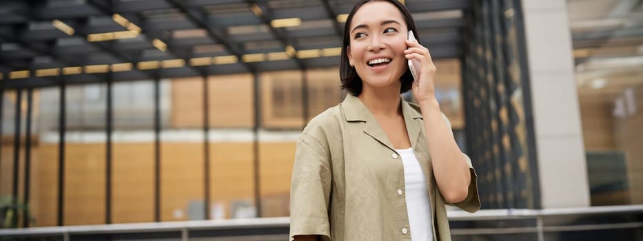 Happy asian woman talking on mobile phone, standing on bus stop in city. Outdoor shot.