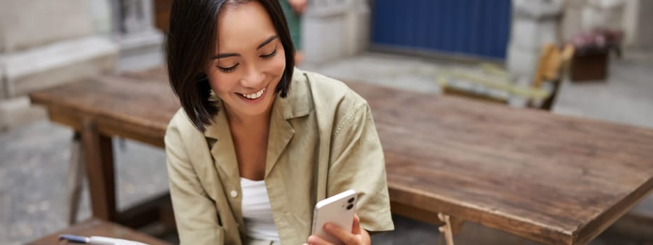 Stylish urban girl sits with her phone in cafe, drinks coffee and chatting, browsing social media on smartphone.