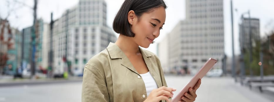 Portrait of asian woman reading, using tablet while standing on street, smiling while looking at screen.