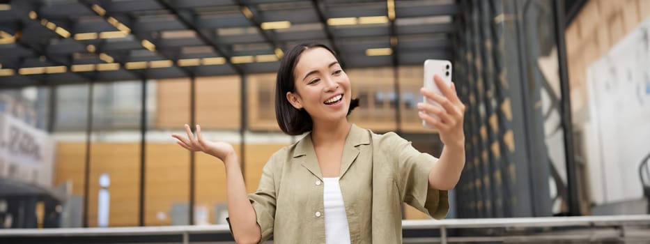 Happy asian girl shows something behind her during video call, demonstrating smth, standing on street.