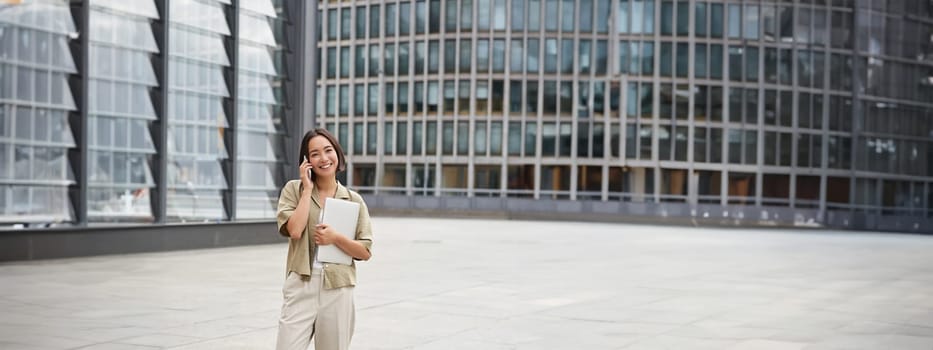 Stylish urban girl walking on street, talking on mobile phone and carrying laptop. Young woman making a telephone call while going somewhere.