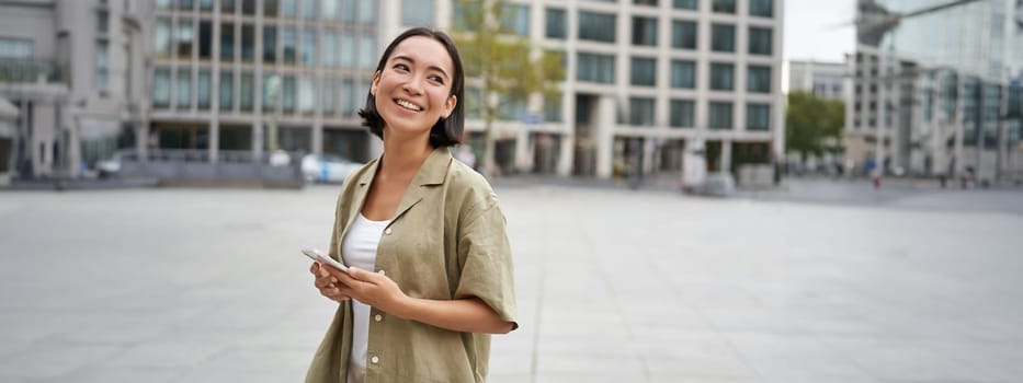 Portrait of asian woman standing on street, city square and holding mobile phone. Girl with smartphone walking outdoors.
