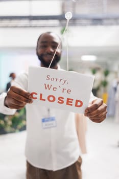 Smiling african american man hanging closed sign in window of fashion boutique, noticing about work day end. Clothes retail shopping mall cheerful assistant putting information sign on door