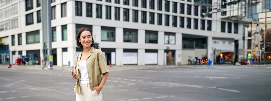 Woman smiling, standing with smartphone on street, waiting for taxi, checking mobile phone app.