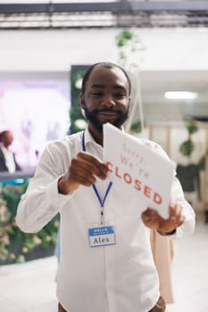 Clothing store smiling african american owner holding sorry we are closed sign. Fashion boutique cheerful man assistant hanging sign informing about end of business hours