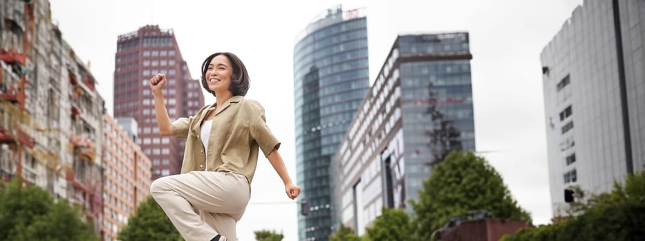 Vertical shot of young asian woman posing happy, raising hands up and dancing, triumphing, celebrating victory, enjoying day out in city.