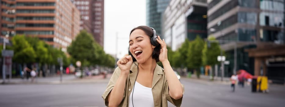 Dancing girl feeling happy in city. Asian woman dancing and listening music in headphones, posing on street.