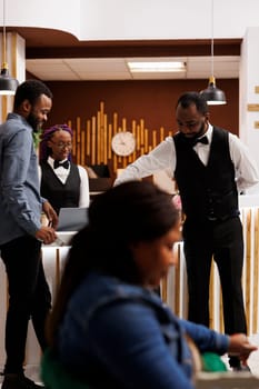 Young African American smiling guy bellboy providing customer service to hotel guest, moving his luggage, carrying suitcase to room after check-in. Bellhop and tourist communicating at front desk