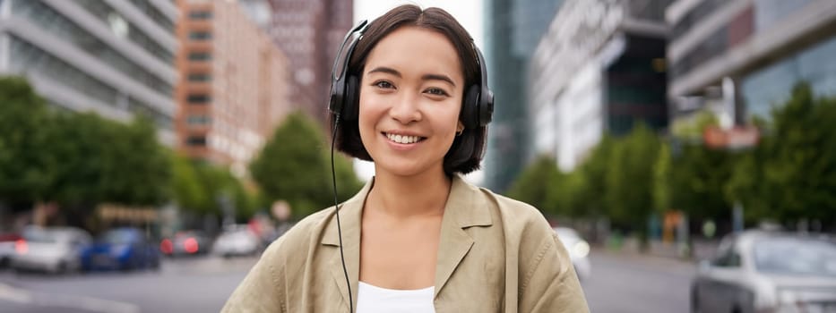 Portrait of smiling asian woman in headphones, standing in city centre on street, looking happy, listening to music.