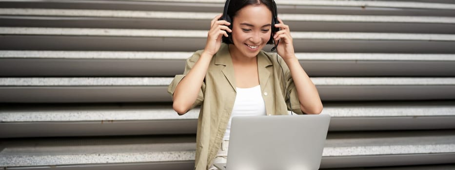 Portrait of asian woman, digital nomad girl using laptop and listens to music outdoors. Young student works on computer and smiles.