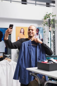 Arab blogger taking selfie on smartphone, creating content for fashion blog in clothing store. Influencer recording video on mobile phone while promoting clothing store offerings