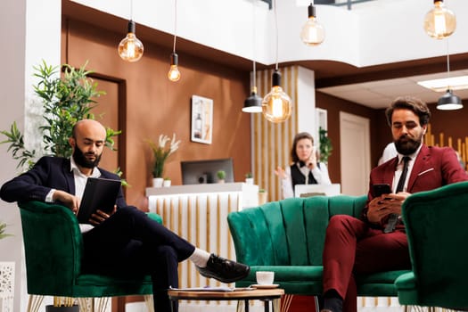 Two businessman in lounge area waiting to do check in process and relax after long flight, travelling on work purposes. White collar workers in hotel lobby preparing for upcoming corporate event.