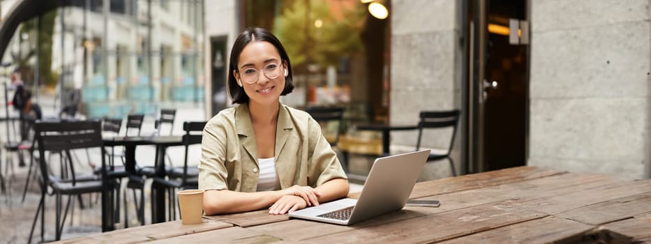 Young woman, student working from cafe, sitting with laptop and cup of coffee outdoors, looking at camera.