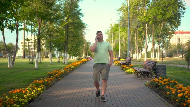 A young man walks in the park and talks on the phone. A man in a polo and shorts is walking along a green alley. 4k