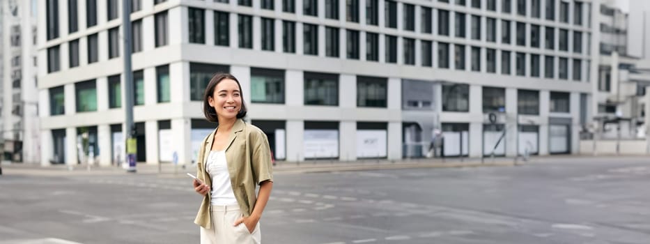 Woman smiling, standing with smartphone on street, waiting for taxi, checking mobile phone app.