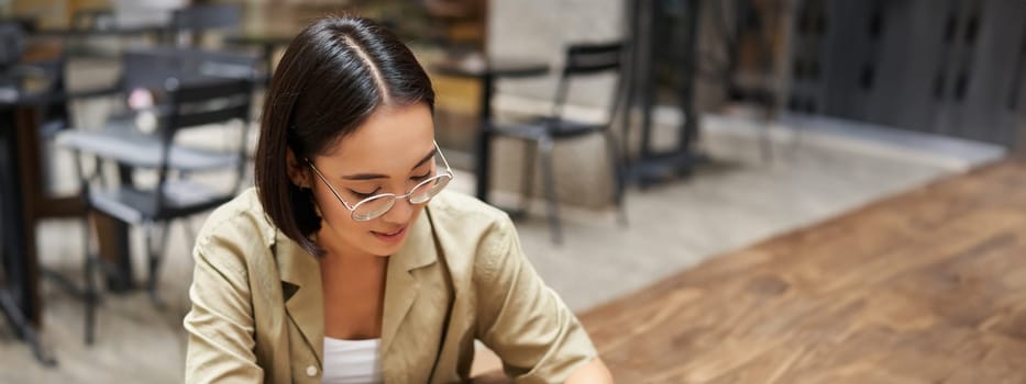 Vertical shot of young asian woman doing homework, making notes, writing something down, sitting in an outdoors cafe and drinking coffee.