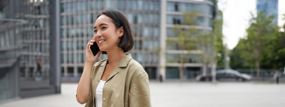 Vertical shot of young asian digital nomad, girl talks on mobile phone and walks on street with laptop. Young woman remote worker going to coworking space.