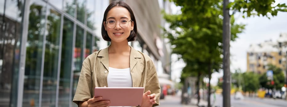 Outdoor shot of young korean woman stands on street with laptop, wears glasses, reads, smiles happily.