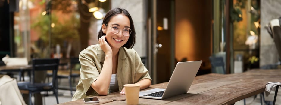 Portrait of smiling girl in glasses, sitting with laptop in outdoor cafe, drinking coffee and working remotely, studying online.