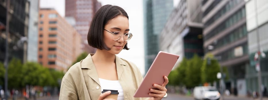 Portrait of happy young woman in glasses, standing on street with cup of coffee and tablet.