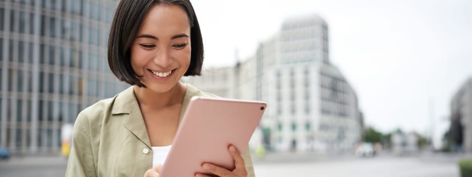 Portrait of asian woman reading, using tablet while standing on street, smiling while looking at screen.