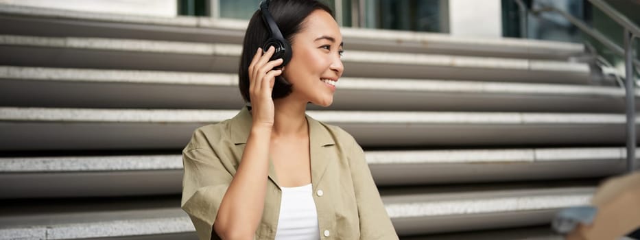 Close up portrait of smiling asian woman listens music in headphones, turns around with happy face expression, resting outdoors.