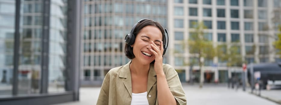 Carefree asian girl, laughing and smiling, wearing headphones and walking on street. Outdoor shot of young woman listening music and looking happy.