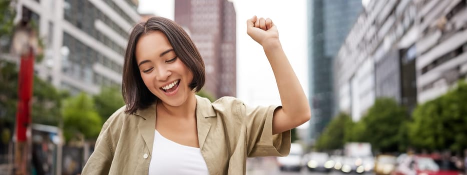 Happy people. Young smiling asian woman dancing in city, raising hands up in city, triumphing, celebrating victory.