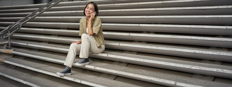 Portrait of asian student girl on stairs talks on mobile phone, smiles at camera, sits outside building.