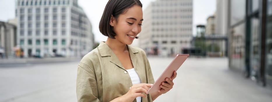 Beautiful girl standing in city square with tablet, reading and drinking takeaway coffee.