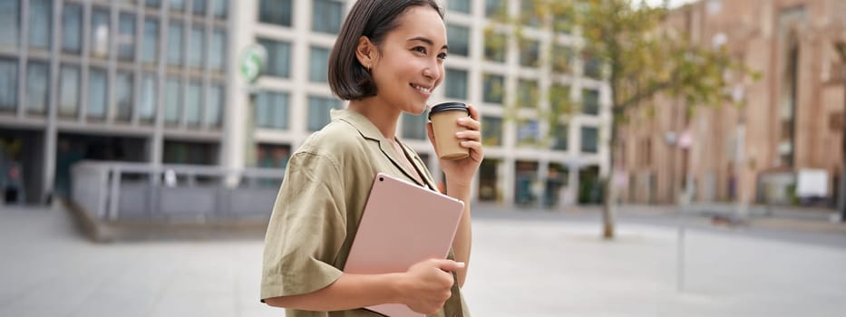 Portrait of asian girl with tablet, drinks coffee on street, walking in city centre.