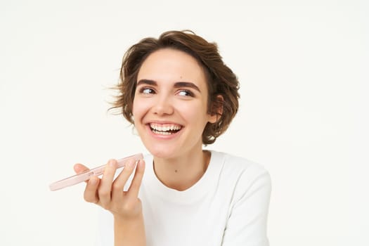 Laughing, happy woman talking on speakerphone, holding phone near mouth, recording voice message on smartphone, standing over white background.