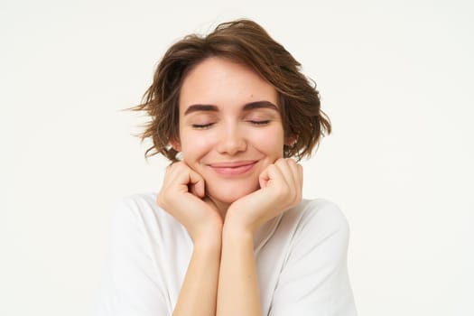 Close up of hopeful young woman, girl anticipating something, touching her face, looking forward to something, waiting with excitement, standing over white background.