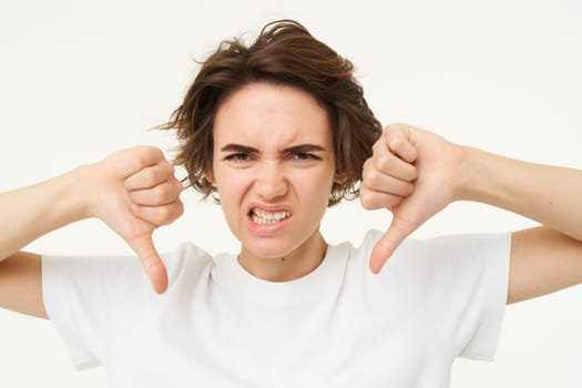 Close up portrait, face of young woman that is showing thumbs down, dislike, do not recommend gesture, express negative feedback, isolated over white background.