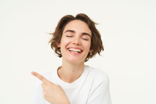 Image of cheerful brunette female model, candid girl points finger left and smiles, shows advertisement, poses over white background.