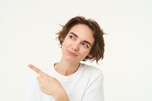 Image of cheerful brunette female model, candid girl points finger left and smiles, shows advertisement, poses over white background.